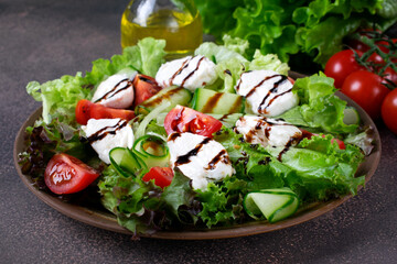 Salad with mozzarella cheese, lettuce, cucumber and tomatoes with spices and olive oil topped with balsamic glaze on brown plate - obrazy, fototapety, plakaty