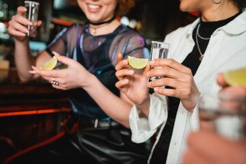 Cropped view of smiling multiethnic women holding tequila and lime in bar.