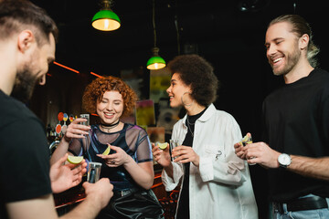 Smiling young interracial women holding tequila and lime near friends in bar at night.