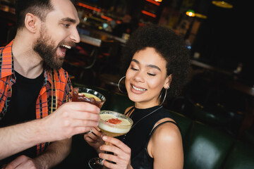 Excited man holding negroni cocktail near curly african american friend in bar.