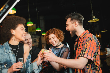 Cheerful friends holding lime and tequila near african american friend in bar.