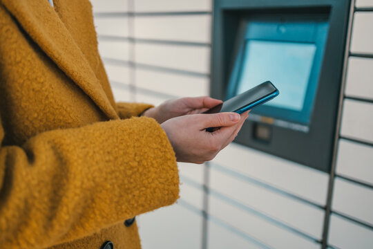 Woman holding smartphone using red automated self service post terminal machine or locker to deposit the parcel for storage. High quality photo