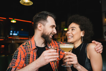 Cheerful man holding cocktail and hugging curly african american friend in bar.