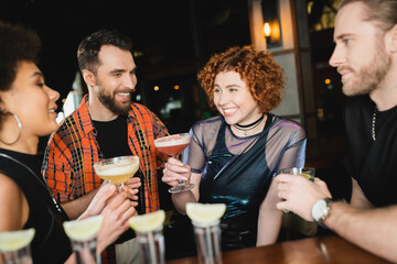 Cheerful woman holding cocktail near multiethnic friends and tequila shots in bar.