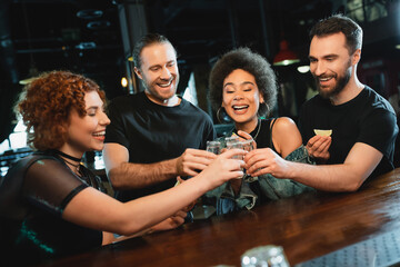 Carefree multiethnic friends toasting with tequila near stand in bar.
