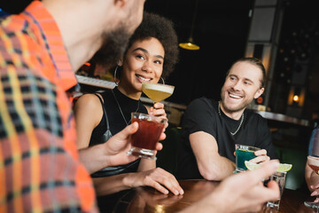 Smiling african american woman holding pisco sour cocktail near friend in bar at night.