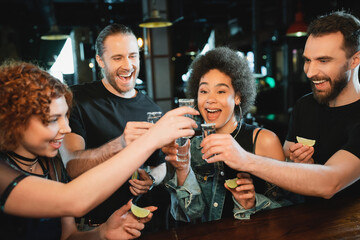Cheerful interracial friends holding tequila and lime near stand in bar. - obrazy, fototapety, plakaty