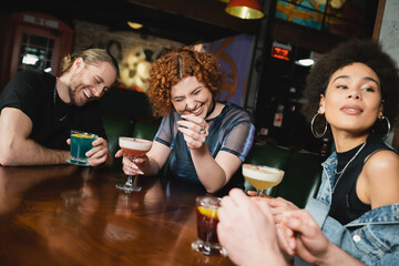 happy interracial friends laughing and holding different cold cocktails in bar.