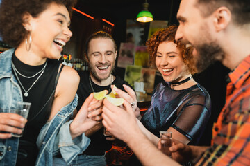 Excited interracial friends holding sour lime and tequila shots in bar.