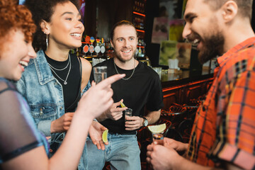 Cheerful interracial friends with tequila shots talking in bar at night.