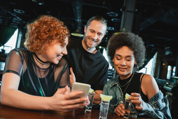 Smiling redhead woman holding smartphone near multiethnic friends and tequila in bar.
