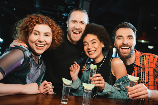 Positive interracial friends holding shots of tequila and looking at camera in bar.