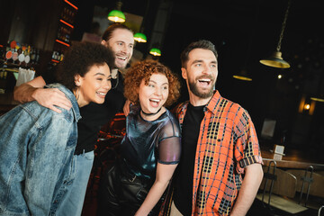 Cheerful interracial friends hugging and looking away in bar at night.