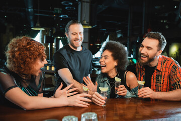 Cheerful interracial friends with tequila talking and laughing near stand in bar.