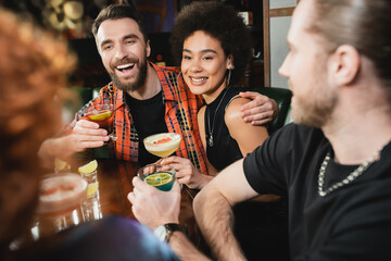 Positive multiethnic people holding different cocktails near blurred friends in bar.