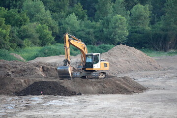 Industrial crawler excavator on top of dirt pile at local road construction site surrounded with gravel and sand with tall uncut grass and dense trees in background