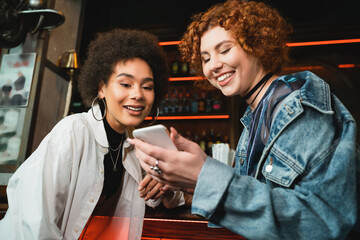 Positive multiethnic friends using smartphone near stand in bar in evening.