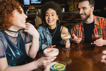 Young curly african american woman holding cocktail near cheerful friends in bar.