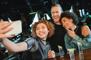 Positive woman taking selfie with multiethnic friends near tequila and lime in bar.