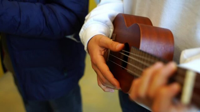 Two children play simple chords on a small wooden ukulele with white and blue hardware and yellowish lights