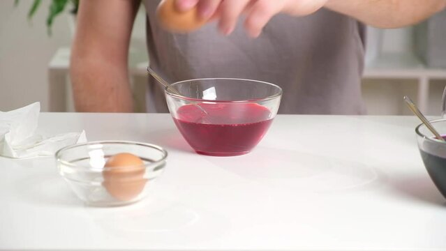 A man puts an egg on a spoon into a glass cup with a dye. Dyeing easter eggs with food coloring is the first step in favorite Easter egg decorating ideas.