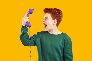 Young boy holding vintage telephone and screaming in it on yellow background. Red-haired guy in a green sweater with open mouth with old purple phone in his hand yelling with anger.