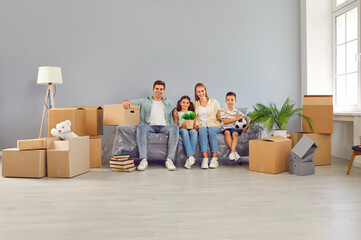 Fototapeta na wymiar Happy friendly family with two children moving in new modern apartment house. Mom, dad and daughter are sitting on couch among boxes, smiling and looking at camera. Moving, family lifestyle concept.