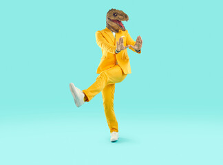 Fototapeta na wymiar Funny man in a dinosaur costume dancing in the studio. Happy guy in a bright yellow suit and funny dinosaur mask dancing on a turquoise blue colour background. Having fun at a party concept