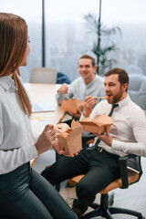 Woman is leaning on the table. Group of coworkers are eating food from eco boxes in the office together