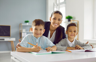 Fototapeta na wymiar Portrait of friendly female teacher with two happy schoolchildren sitting together at desk. Teacher helps and advises two schoolboys on school assignment. Back to school concept.