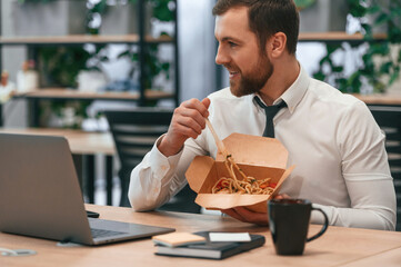 Sitting by laptop. Businessman in formal clothes is in the office eating udon that is in the eco box