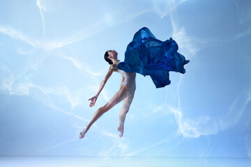 Light as feather. Shot of beautiful jumping ballerina with silk fabric over blue background