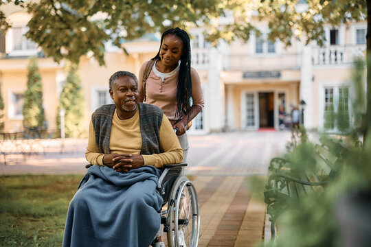 Happy black woman pushes her senior father in wheelchair while visiting his at nursing home.