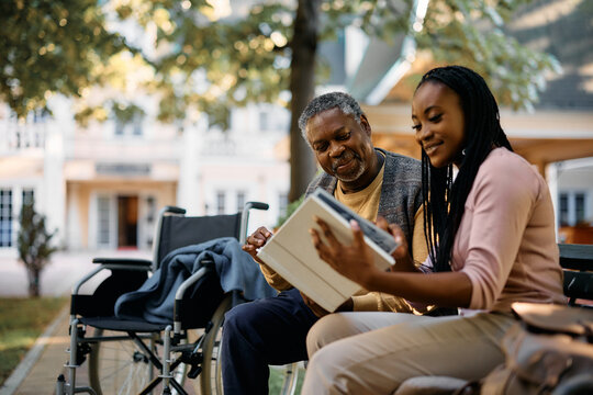 Happy black woman looking at photos in family album with her senior father while vising him at nursing home.
