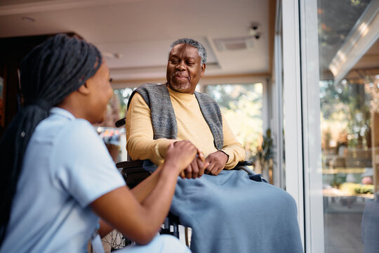 Smiling black senior man in wheelchair talks to nurse while holding her hand in nursing home.