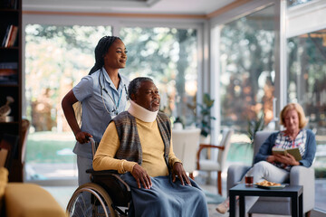 Young black nurse pushing senior man in wheelchair through nursing home.