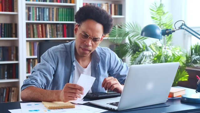Young focused smart African American man auditor enters data on financial expenses into laptop by retyping amounts from cashier checks into CPM system sits at desk in home office. Accounting, finance