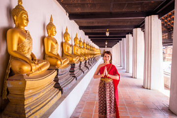 Young woman dressed in traditional red Thai dress and golden ornaments stands sawasdee as a welcome...