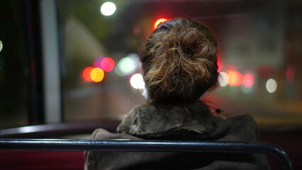 Back of female commuter riding bus at night after work. Passenger sitting inside public...