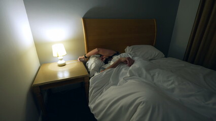 Obraz premium Man laying down in bed to rest after long day. Person going to sleep going under bedsheets resting turning bedside lamp off