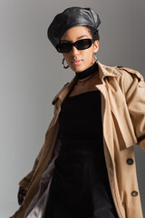 Trendy african american model in sunglasses and trench coat standing isolated on grey.