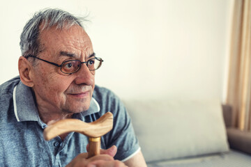 Shot of a senior man sitting alone on the sofa at home and looking contemplative while holding his walking stick.