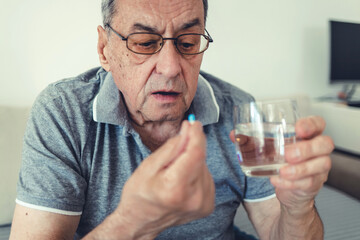 Elderly man taking pills for depression sitting on couch. Old upset patient swallowing pill with glass of water.