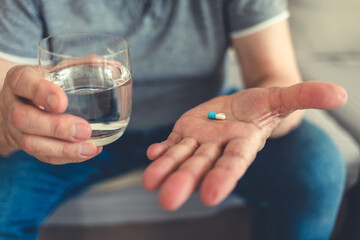 Elderly man taking pills for depression sitting on couch. Old upset patient swallowing pill with glass of water.
