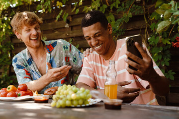 Cheerful male friends laughing while using mobile phone during barbeque party