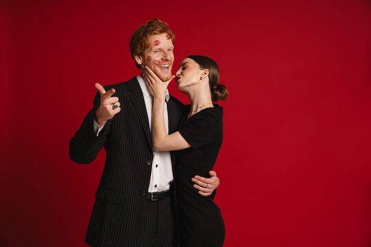 Cheerful man with red lipstick marks on his face pointing finger at camera while woman kissing him isolated