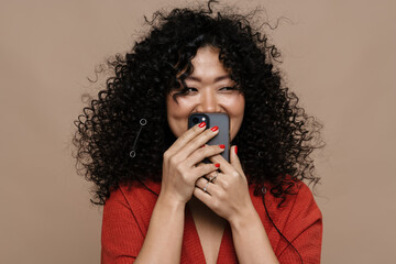 Ceerful asian woman covering her face with mobile phone isolated over beige background
