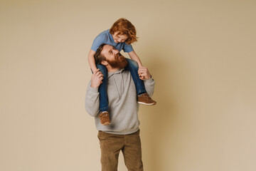 Father smiling while carrying son on his shoulders isolated over beige wall