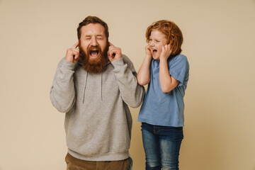 Fototapeta premium Father and son covering their ears with fingers and screaming while standing isolated over beige background