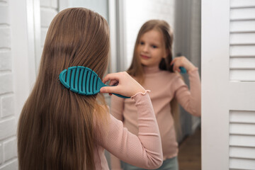 Cute teenager girl child combing her long blonde hair and smiling looking in the mirror, view from the back, Selective focus.